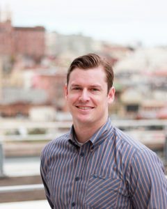 Headshot image of Alex in front of a blurry urban background